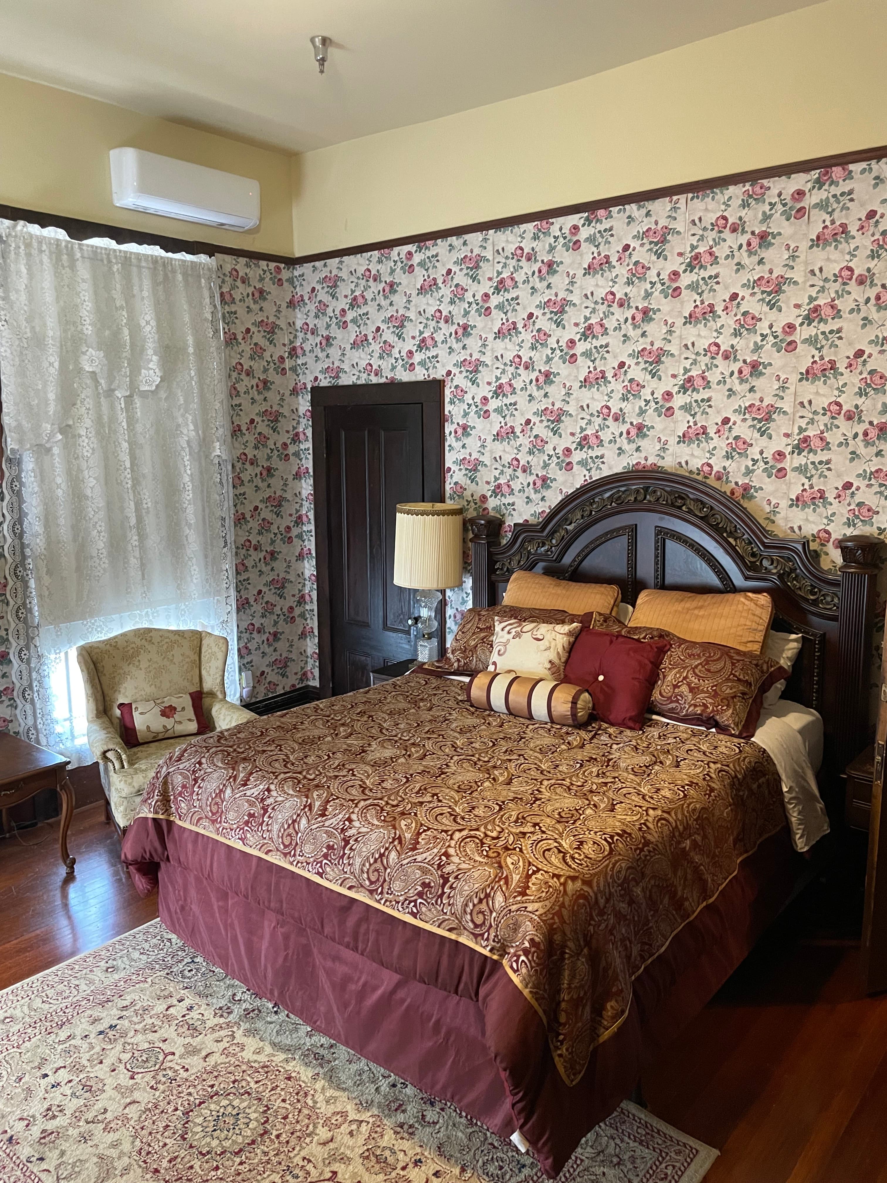 A cozy bedroom featuring a large ornate bed, floral wallpaper, a lace-curtained window, and a vintage armchair.